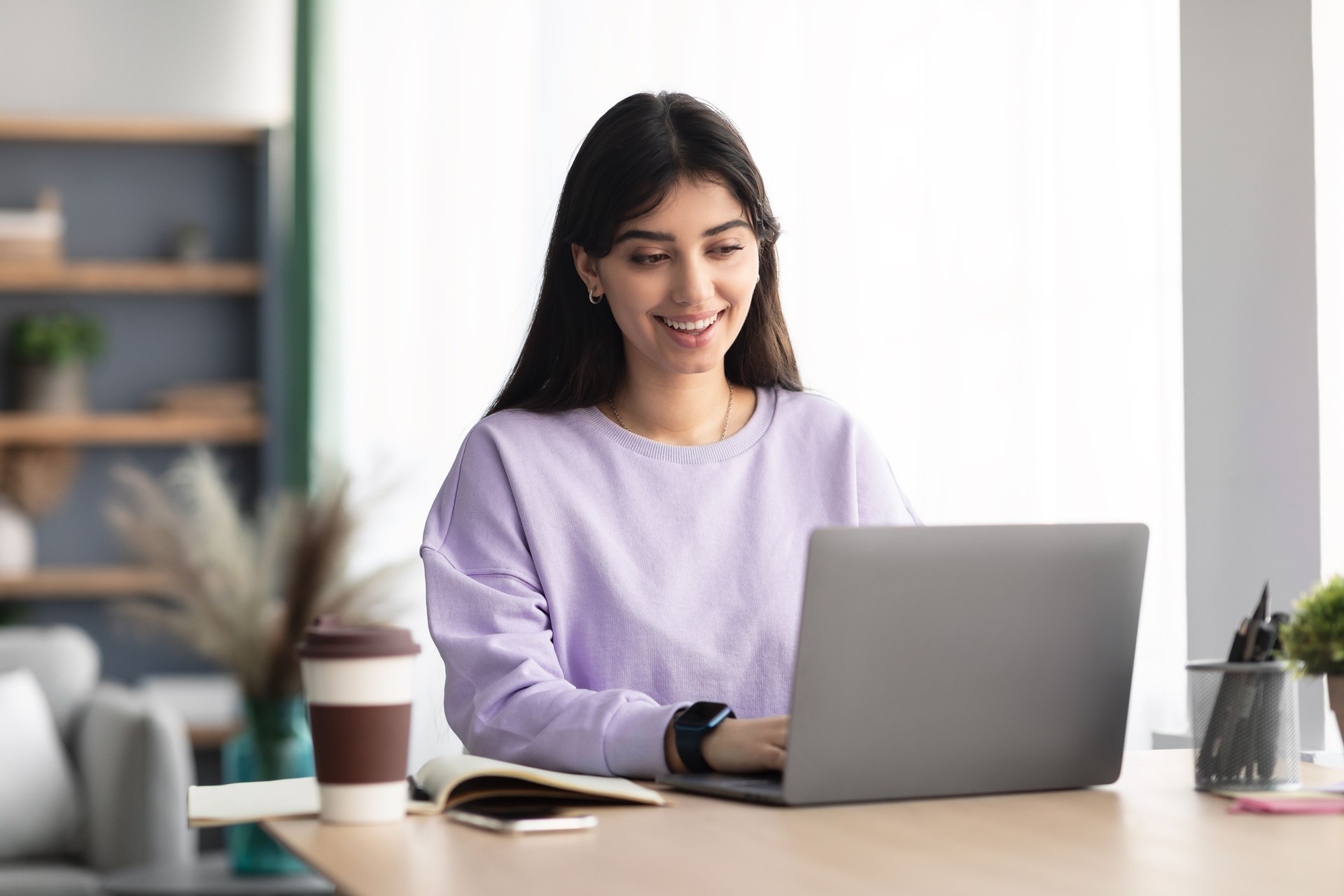 Cheerful millennial lady sitting at desk using laptop